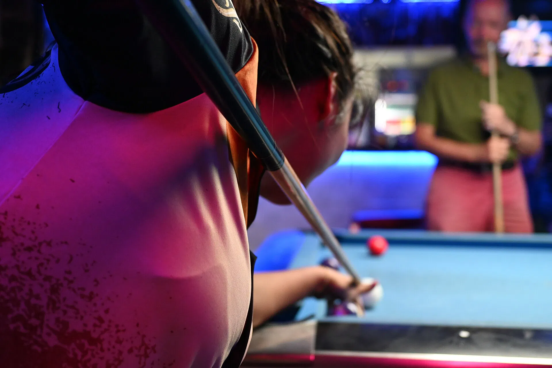 A pool player lines up a shot under colorful bar lights while the opponent watches from the background